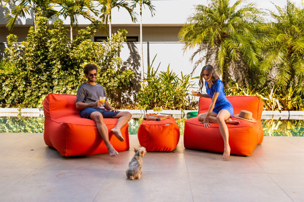 Casal relaxa em puffs Paradiso junto à piscina, rindo e confortável. Um cachorro brinca próximo. Ambiente externo aconchegante